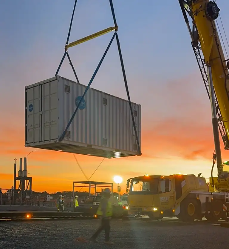 HydrogenCube 20 container being lifted into position by a crane at an industrial site during sunset, with workers and heavy equipment visible in the foreground.