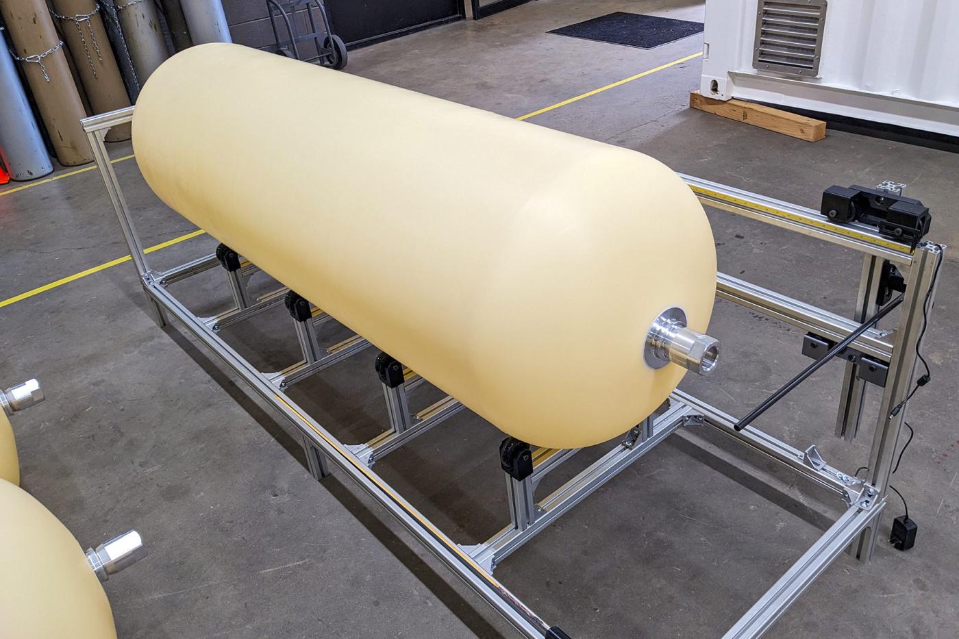 Type IV polymer liner mounted on a metal support frame during the Advanced Liner Fabrication process, showing its smooth cylindrical shape with rounded ends and metallic fittings.